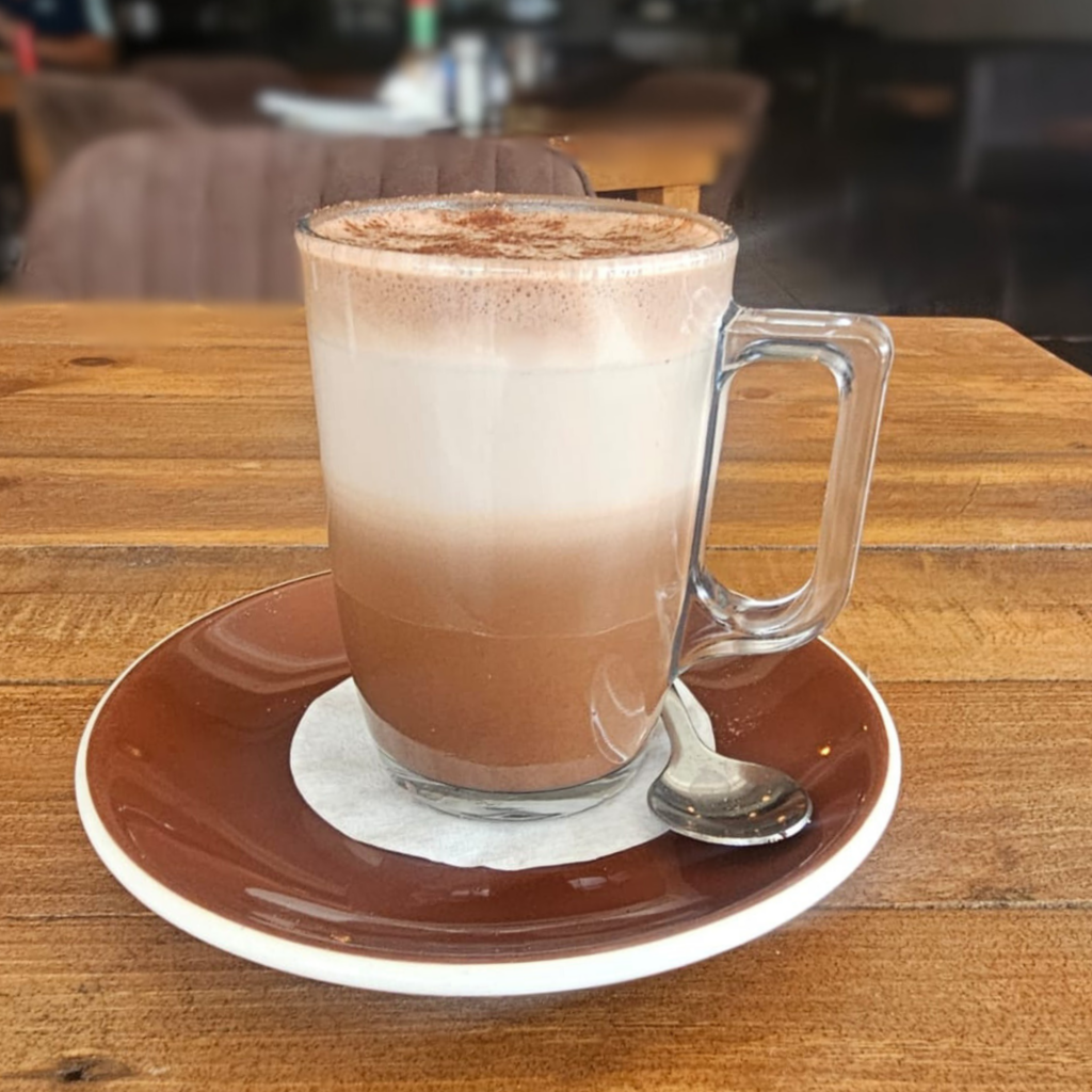 Creamy layered mocha in a clear glass mug topped with frothy milk and cocoa on a wooden table at My Hometown Café Newton Park